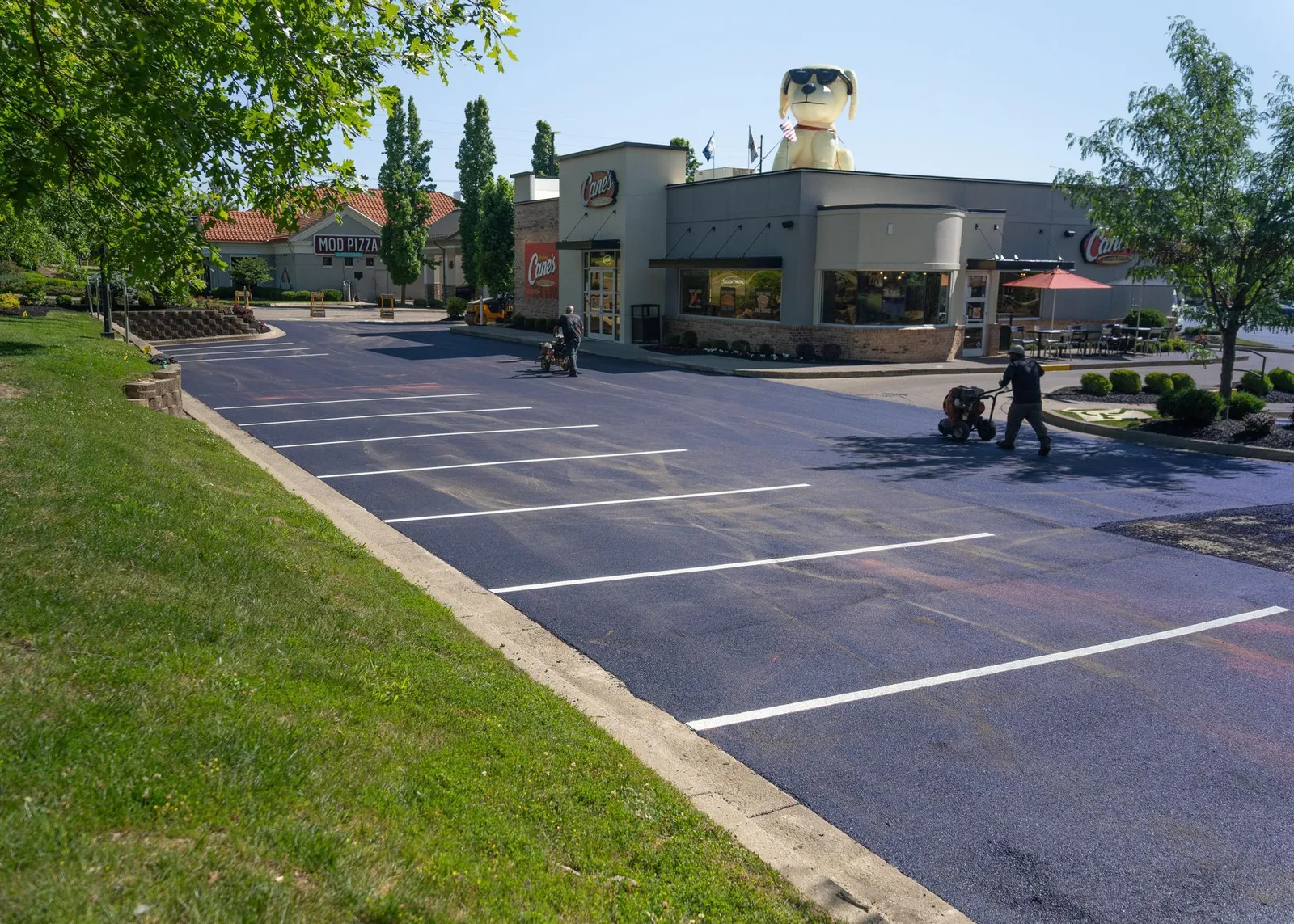 Parking lot beside a small building with a tree-lined lawn and two people walking near the entrance