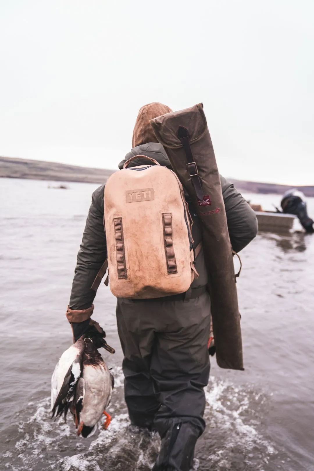A waterfowl hunter wading into a lake carrying a gun case and backpack