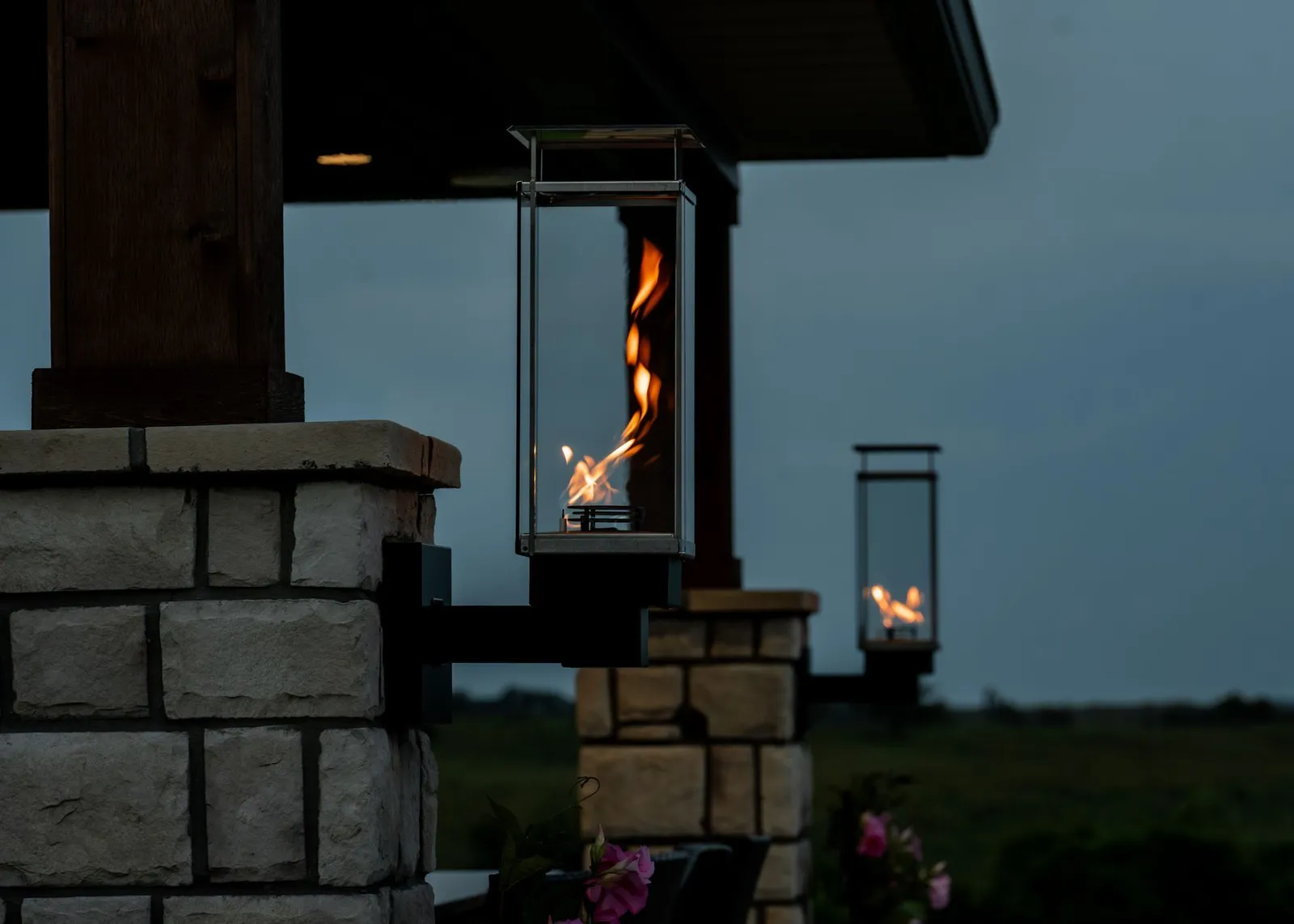 Lit lanterns hanging from a stone pillar at dusk, with one flame glowing in the foreground and another in the distance