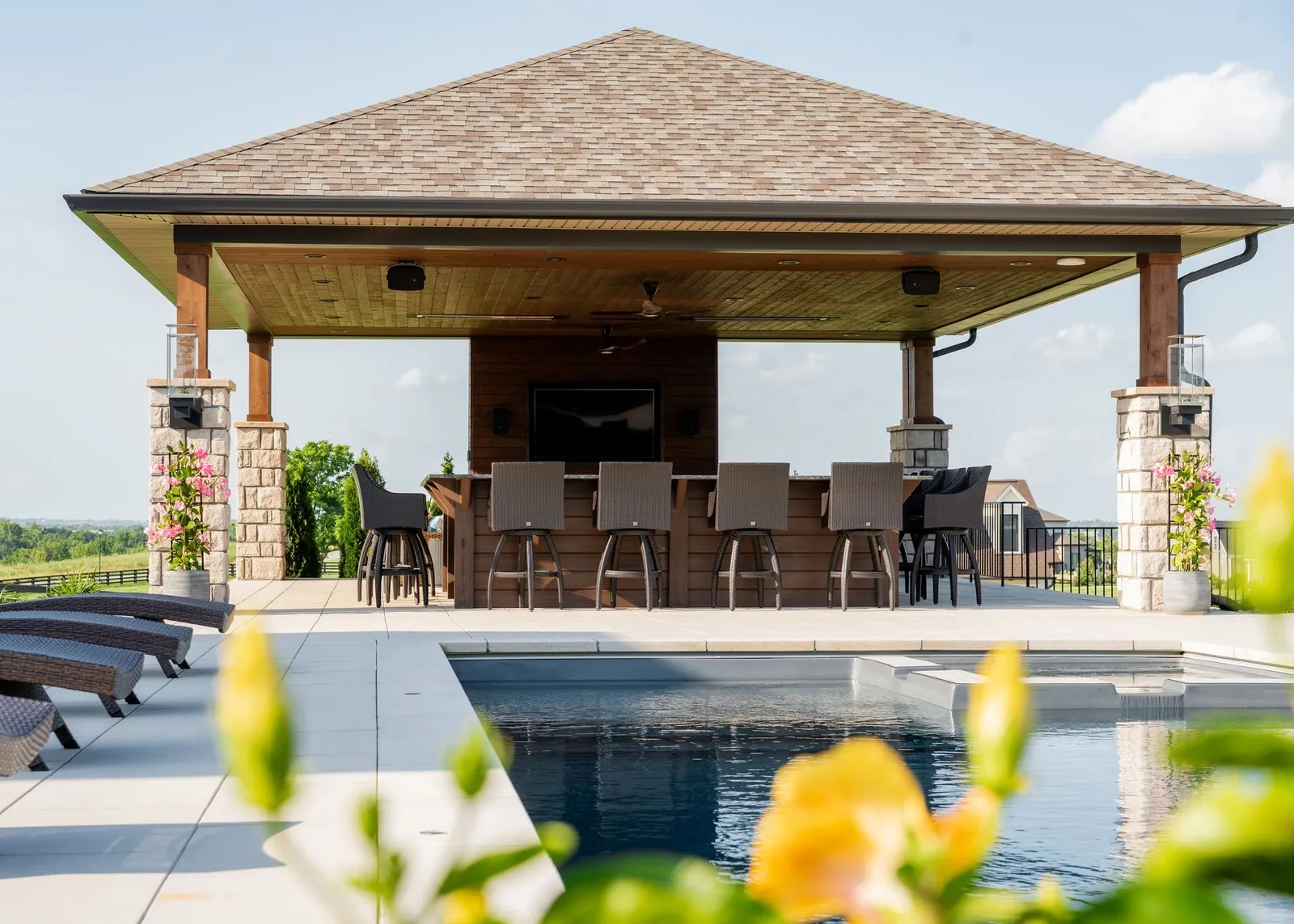 Poolside patio with a thatched-roof pavilion, dining table, and lounge chairs overlooking a bright landscape