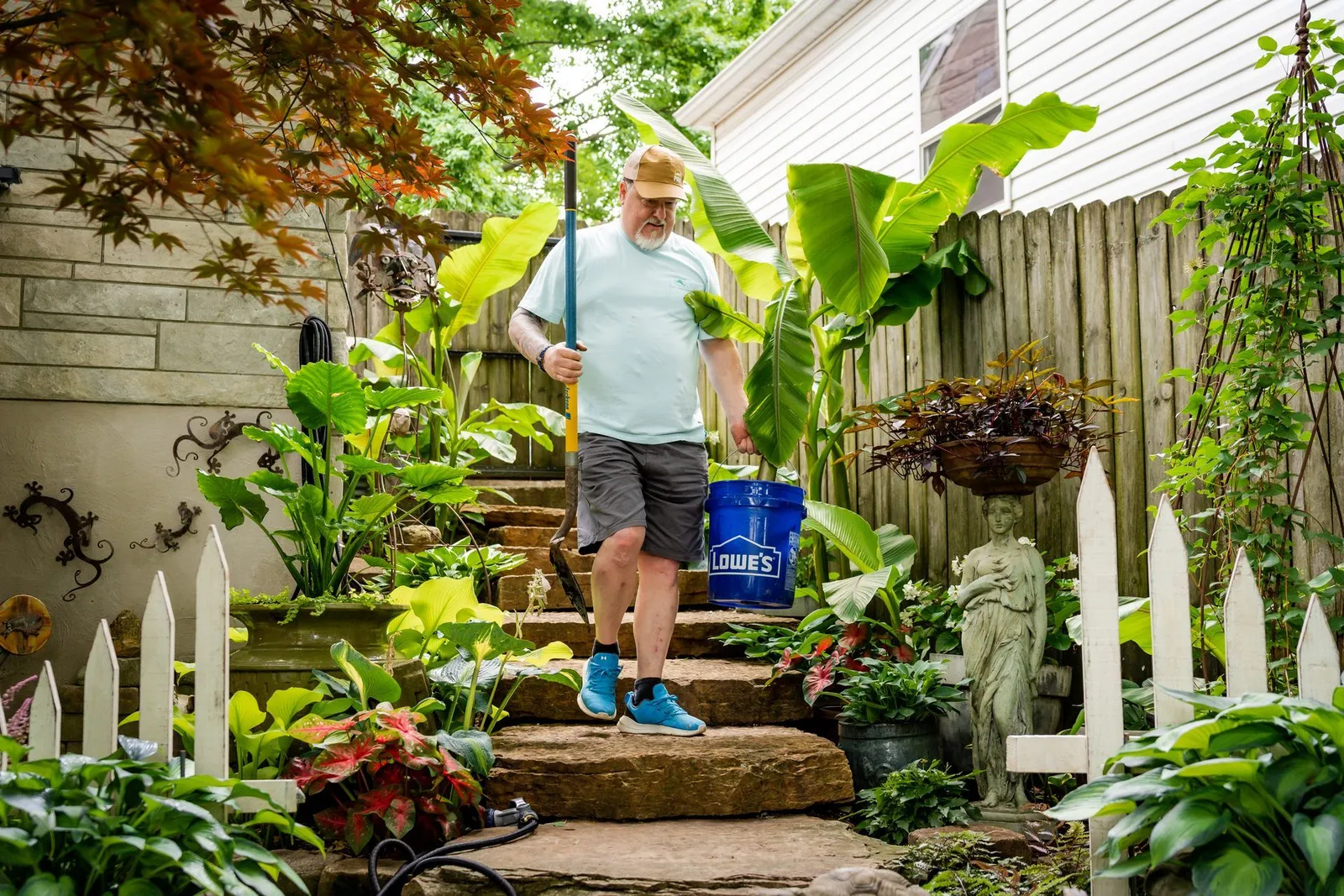 A gardener carrying a blue bucket up stone steps through a backyard garden