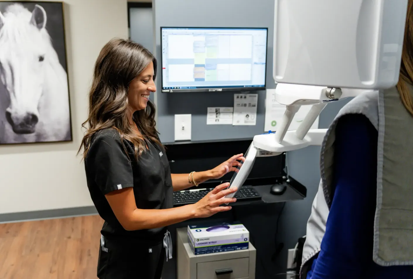 Technician operating a medical imaging machine beside a horse photo in a clinic room